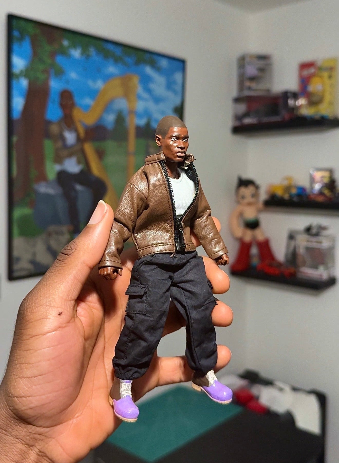 A hand holding a small action figure of a man in a brown jacket, white shirt, and dark pants, with a blurred room in the background containing shelves and a framed picture.
