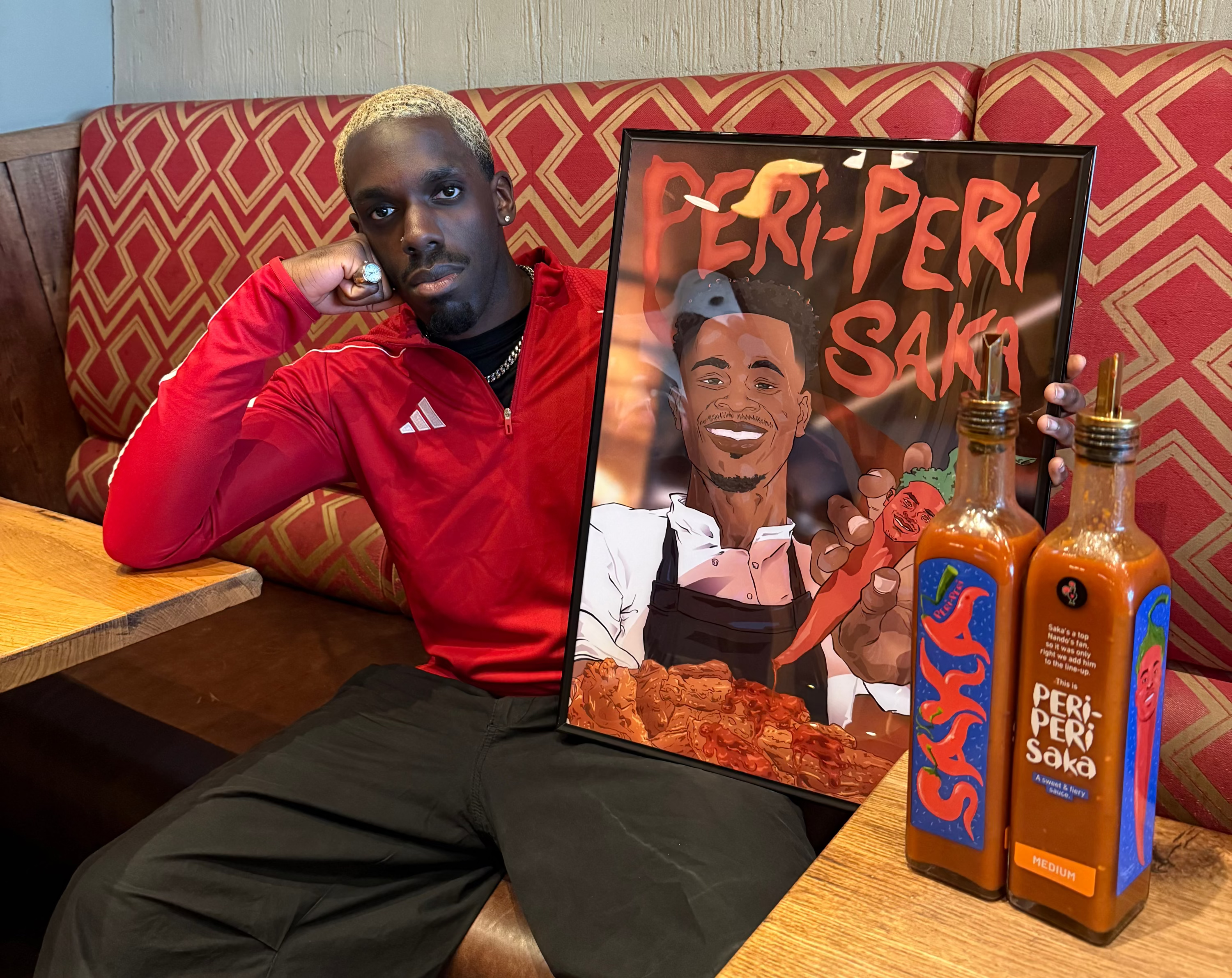 A man in a red Adidas sweatshirt and black pants sits in a restaurant booth next to a large framed poster reading 'FERI-PERI SAKE' and two bottles of Feri-Peri Sake sauce on the table.