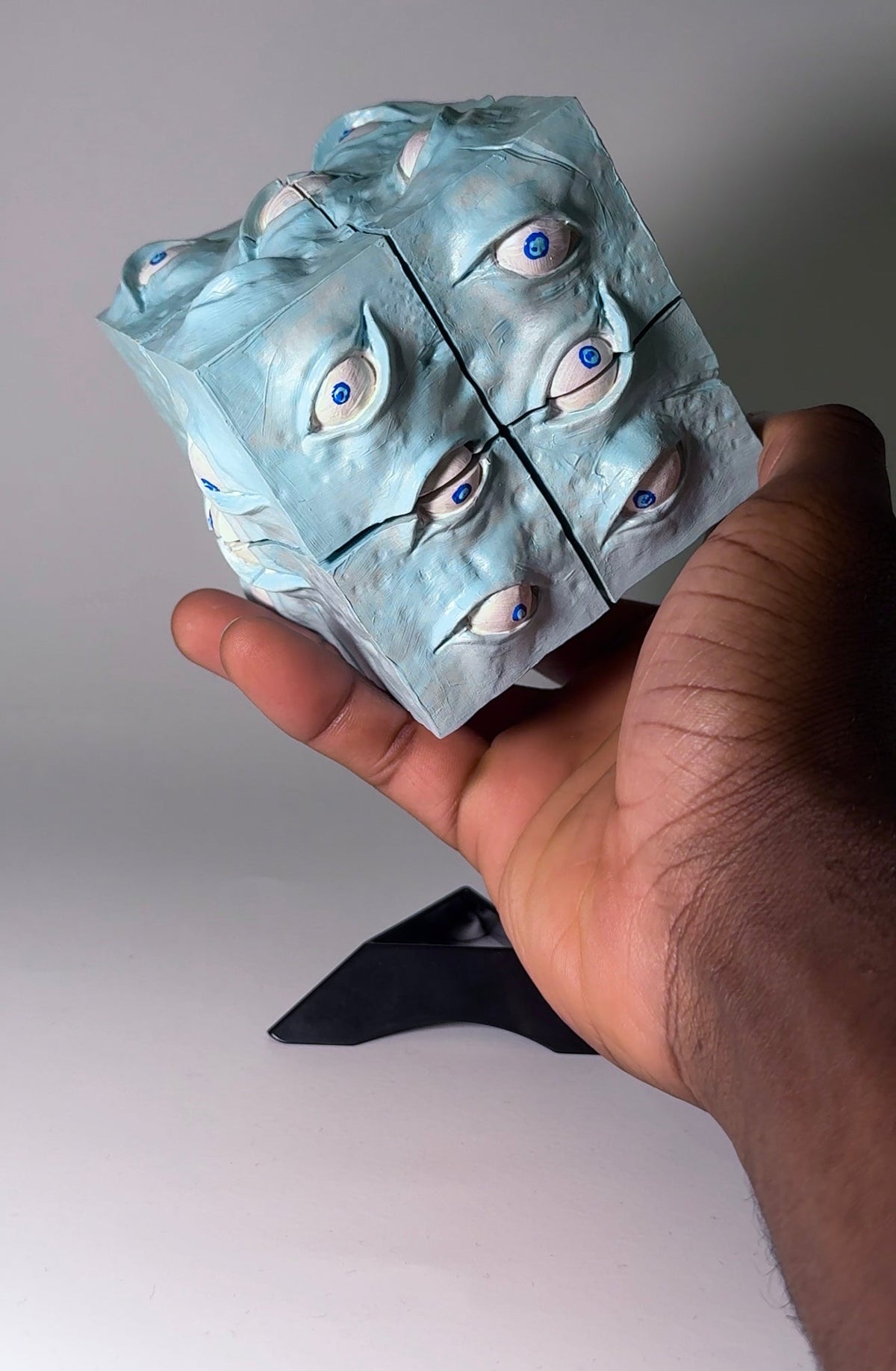 A hand holding a 3D-printed optical illusion cube that appears to be made of multiple faces of the same eye design in light blue, white, and gray, with black lines separating the sections.
