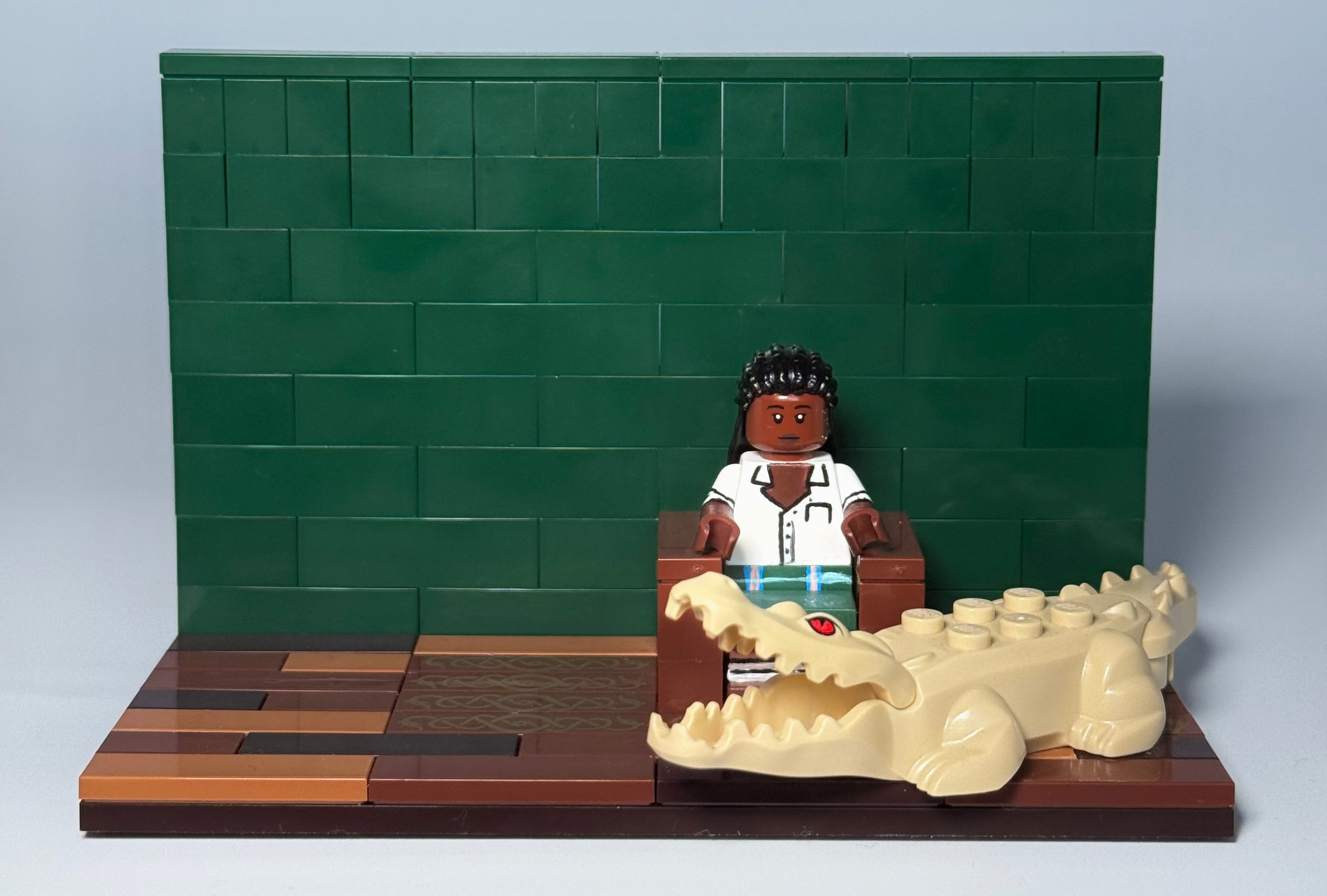 A miniature LEGO-style doctor scene with a doctor minifigure seated at a desk in front of a green brick wall, alongside a beige alligator figure on a wooden base.